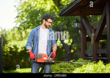 Jardinier masculin professionnel dans des lunettes de protection façonnant des haies denses avec des coupe-branches électriques sur la cour arrière. Vue de face du gars dans la chemise élaguant arbuste avec coupe-herbe à la journée ensoleillée. Concept de hobby, travail. Banque D'Images