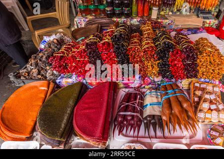 Cuir de fruits (peut-être tklapi, géorgien ტყლაპი) repose en grandes feuilles rondes orange, vert et rouge devant un étal de churchkhela au Dezerter Bazaar (Marché des déserteurs) à Tbilissi, Géorgie. À côté se trouvent des brins de churchkhela enveloppés dans du film avec des rubans bleus et des sacs étiquetés dans un panier. Derrière eux se trouvent des bouquets de morceaux de fruits brillants enfilés, en rouge, noir et orange, enveloppés de rubans dorés. Des fruits séchés enfilés sur des cordes (peut-être des persimmons) reposent à gauche. Le tklapi est un cuir de fruits traditionnel géorgien fait à partir de purée de fruits. Le tklapi acide est fait à partir de prunes cerises, sucré Banque D'Images