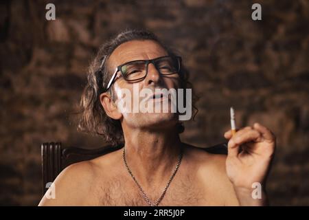 Un homme plus âgé, âgé d'environ 60 ans, de nationalité serbe, aux cheveux longs et aux lunettes, fume pensivement une cigarette sur fond sombre. Artiste, CRE Banque D'Images