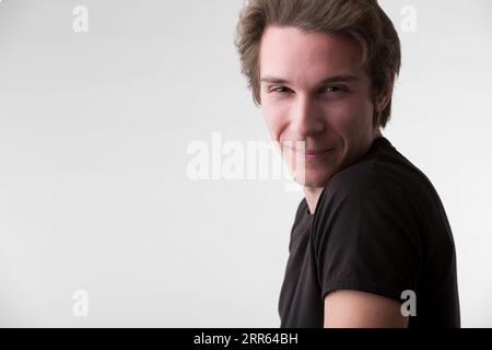 Sur un fond neutre se dresse un jeune homme attrayant dans un tee-shirt noir, ses cheveux bruns courts. Ses yeux intenses et son sourire suggèrent des opinions profondes et skil Banque D'Images