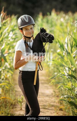 Un portrait d'une fille - un jockey dans un casque et un T-shirt blanc, qui tient un cheval noir dans ses mains - un jouet sur un bâton, se tient dans le maïs. Hobbyhor Banque D'Images