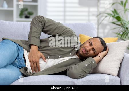 Jeune homme afro-américain allongé sur un canapé à la maison grimaçant et souffrant de douleurs, tenant le ventre avec les mains. Gros plan. Banque D'Images