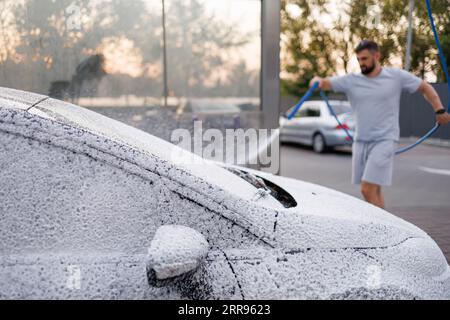 Le capot et le pare-brise de la voiture sont recouverts de mousse, l'homme en arrière-plan applique de la mousse sur la voiture. Une voiture dans un lave-auto en libre-service. Banque D'Images