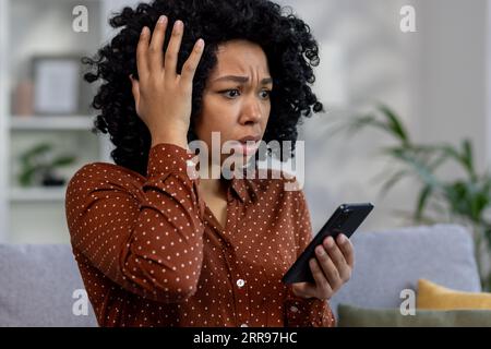 Femme afro-américaine inquiète assise sur le canapé à la maison, regardant l'écran du téléphone et tenant la tête en état de choc. Banque D'Images