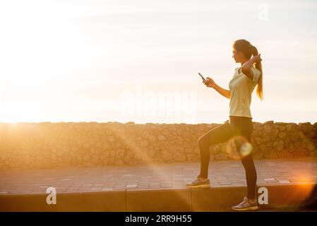 Portrait rapproché d'une jeune femme en vêtements de sport, tenant son smartphone, emmène le selfie entre les entraînements sur fond d'un ciel orange à l'aube. Le c Banque D'Images