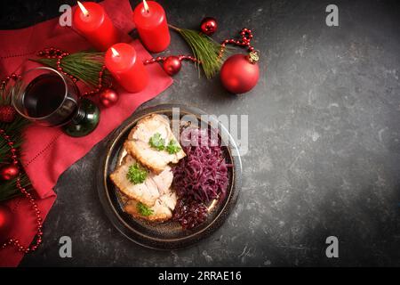 Dîner de Noël, tranches de porc rôti avec du chou rouge sur une assiette, bougies rouges, vin, serviette et décoration sur une table sombre, vue à angle élevé d'en haut, Banque D'Images