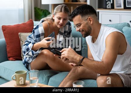 Couple amoureux assis sur le canapé à la maison. Homme et femme se relaxant sur le canapé avec smartphone dans le salon. Les jeunes se détendent dans le temps libre en s'amusant W Banque D'Images