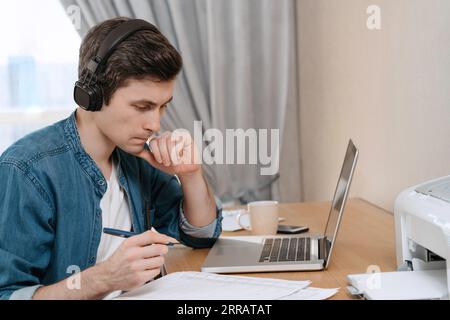 Travailleur sérieux homme freelance dans un casque regardant concentré sur les documents et tenant un pan. Jeune homme d'affaires utilisant un logiciel portable pour les tâches de travail, pensant à la solution des problèmes de travail Banque D'Images