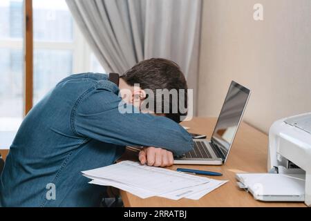 Burnout jeune homme pleure dans son bureau à domicile, assis à son bureau avec un ordinateur portable et des documents. Freelancer déprimé fatigué frustré par l'échec d'entreprise, se sentir épuisé, inquiet des problèmes de travail Banque D'Images