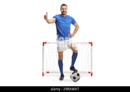 Joueur de football en maillot bleu avec sa jambe sur une balle montrant le pouce devant un mini but isolé sur fond blanc Banque D'Images