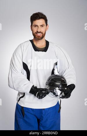 Heureux joueur de hockey sur glace avec casque noir posant à la caméra après match en studio. Portrait de sportif masculin en pleine vitesse, souriant à la caméra, isolé sur fond gris. Concept de sports d'hiver. Banque D'Images