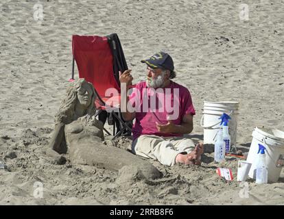 Un artiste de plage âgé est assis dans le sable à côté de sa création de sirène le long de la Promenade à Seaside, Oregon. Banque D'Images