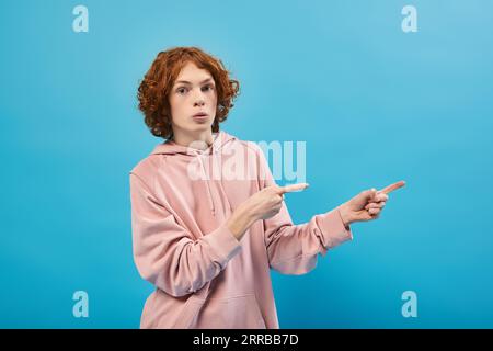 surpris adolescent roux dans un sweat à capuche tendance regardant la caméra et pointant vers le loin avec les doigts sur le bleu Banque D'Images