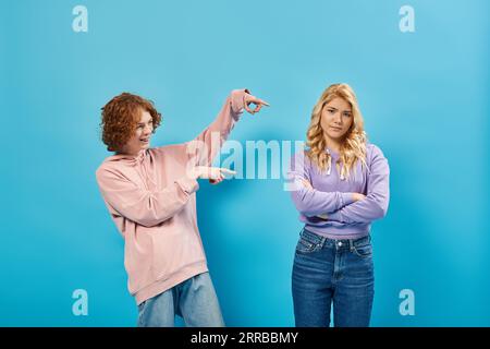 riant rousse adolescent pointant du doigt la petite amie offensée debout avec les bras pliés sur bleu Banque D'Images