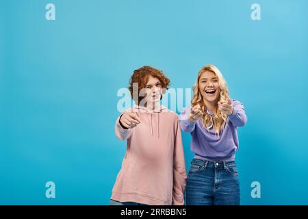 amis adolescents joyeux et à la mode dans des sweats à capuche pointant du doigt vers la caméra et riant sur le bleu Banque D'Images