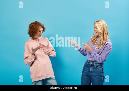 adolescente blonde riant et pointant du doigt vers le petit ami roux découragé sur bleu Banque D'Images