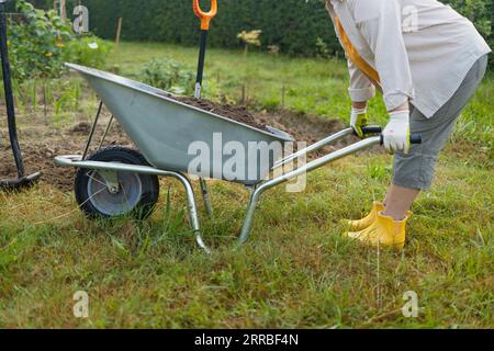 femme fermière senior des années 50 avec une pelle décharge une brouette remplie de terre. Travaux sur le terrain à ressort Banque D'Images