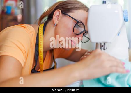 Femme couturière dans les lunettes travaille sur la machine à coudre à son lieu de travail dans l'atelier Banque D'Images