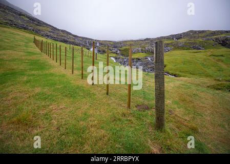 Des cascades d'eau descendent une pente au-dessus du village de Tjørnuvik dans les îles Féroé, avec des clôtures séchant l'herbe au premier plan. Banque D'Images