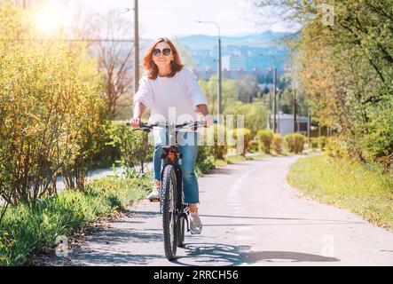 Une femme caucasienne souriante heureuse dans des lunettes de soleil fantaisie chevauchant un vélo sur la rue du parc de la ville. Écologie transport durable et personnes heureuses concept i Banque D'Images
