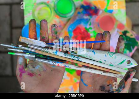 Mains de l'enfant tachées de peinture multicolore après avoir peint avec des pinceaux et un couteau à palette gros plan. Banque D'Images