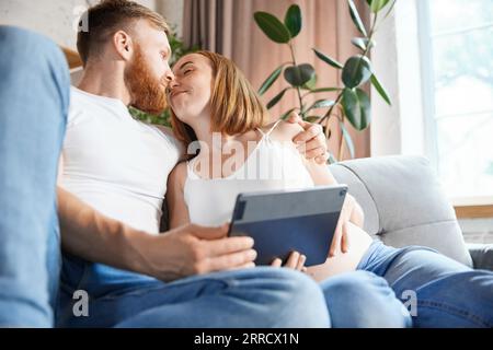 Baisers et amour. Couple, parents assis à la maison et regardant sur la tablette. Attendre bébé, apprendre des informations Banque D'Images