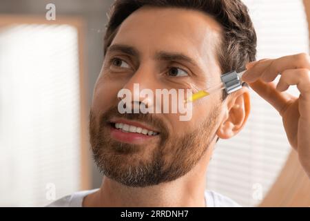 Bel homme appliquant du sérum cosmétique sur son visage à l'intérieur, gros plan Banque D'Images