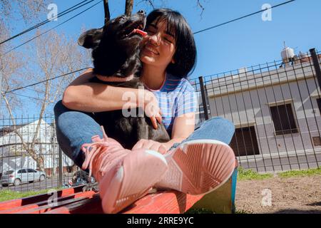heureuse jeune femme latine aux cheveux noirs assise sur un banc avec son chien souriant et jouant dans le parc au coucher du soleil, profitant de la journée, espace de copie. Banque D'Images