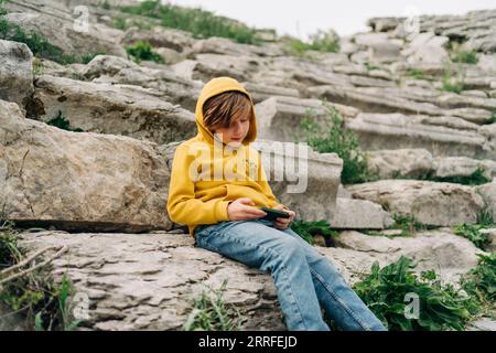 Enfant d'école jouant avec son téléphone portable smartphone dans un ancien amphithéâtre en pierre. Garçon à capuchon jaune en envoyant un SMS au-dessus de son gadget de téléphone pendant Banque D'Images