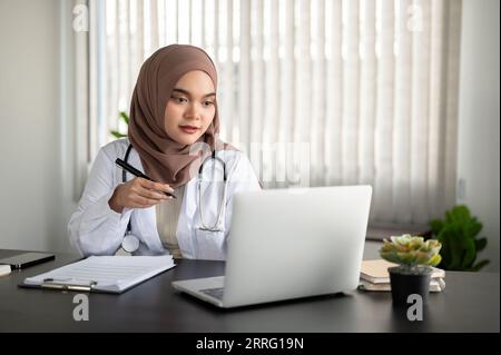 Une belle et professionnelle femme médecin musulmane asiatique en uniforme et hijab travaille sur son ordinateur portable à son bureau dans le bureau d'un hôpital ou cli Banque D'Images