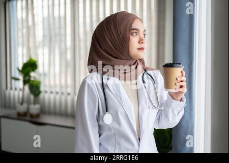 Un portrait d'une femme médecin musulmane asiatique belle et déterminée dans un uniforme et hijab regarde par la fenêtre avec un visage réfléchi tout en havi Banque D'Images