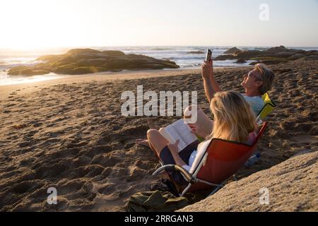 Couple mature assis dans des chaises sur la plage au coucher du soleil avec tablette et livre Banque D'Images