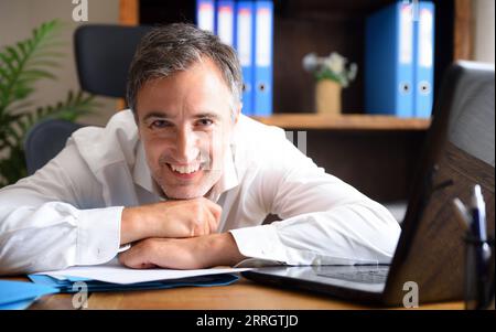 Homme d'affaires entrepreneur en chemise blanche souriant et s'appuyant sur une table de bureau avec des documents et un ordinateur portable et des étagères avec des dossiers en arrière-plan Banque D'Images