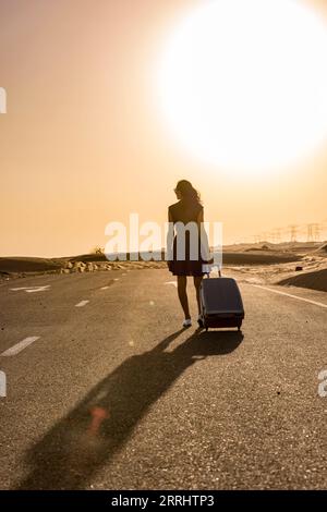 Une femme marche le long d'une route rurale ensoleillée dans le vaste paysage désertique, transportant ses bagages, capturés à contre-jour, avec une lumière chaude. Banque D'Images