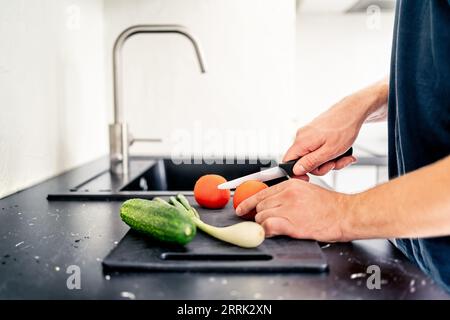 Couper la salade avec un couteau bien aiguisé. Homme cuisinant. Hacher les tomates et trancher les légumes. Préparez la nourriture dans la cuisine à la maison. Repas végétarien sain. Banque D'Images