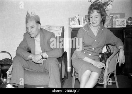 Angleterre. c.1960 – Un homme et une femme sont assis dans un salon à regarder la télévision le jour de Noël. L'homme porte un costume en tweed avec un pull et un chapeau en papier de soie d'un craquelin de Noël. Il est assis dans un fauteuil avec un journal drapé sur le dos. La femme porte un costume deux pièces et une ficelle de perles et est assise dans une chaise de type Lloyd Loom. Derrière eux se trouve un buffet en bois sombre avec des cartes de voeux affichées sur le dessus. Sur le côté se trouve une chauve-souris de charbon en laiton et des fers à feu. Banque D'Images