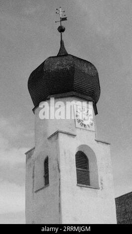 Tyrol, Autriche. c.1960 – l'horloge/le clocher de St. Église Antoine de Padoue située sur Dorfstraße à St Anton am Arlberg. La tour a un toit en dôme en « oignon » carrelé surmonté de la Croix de Lorraine avec girouette intégrée. La tour a une horloge avec des fenêtres à persiennes ci-dessous. Banque D'Images