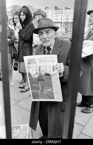 Londres. 1965 – un homme âgé portant un chapeau trilby et un pardessus se tient sur le trottoir à Cumberland Gate, près de Marble Arch, à Londres. Il regarde à travers les balustrades du photographe, qui se tient à Hyde Park, près de Speaker’s Corner. L'homme vend le journal Soviet Weekly et détient un exemplaire daté du samedi 15 mai 1965. Derrière lui se trouve un autre homme qui vend des exemplaires du journal Labour Worker. Un groupe de jeunes femmes sont rassemblés sur le trottoir regardant. Banque D'Images