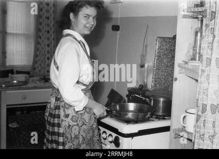 Allemagne de l'Ouest. 1959 – Une jeune femme, portant un tablier, se tient debout devant une cuisinière à gaz dans une cuisine, cuisinant un repas dans une poêle à frire. Elle regarde le photographe et sourit. À côté du four se trouve une étagère en bois recouverte d'un rideau décoré d'un design moderne de style « âge atomique », populaire à l'époque. Banque D'Images