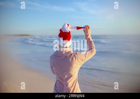 Vu de derrière femme moderne d'âge moyen dans un pull confortable avec chapeau de noël rayé ayant du temps amusant à la plage dans la soirée. Banque D'Images