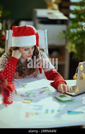 Noël. Femme de propriétaire de petite entreprise élégante stressée dans le chapeau de père Noël et pull de Noël rouge avec des documents et un ordinateur portable travaillant dans le vert moderne o Banque D'Images