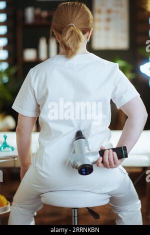 Le temps des soins de santé. Vu de derrière la femme thérapeute de massage dans le salon de spa avec pistolet de massage. Banque D'Images