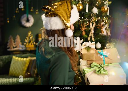 Noël. Vu de derrière femme au foyer moderne d'âge moyen avec chapeau de Père Noël et boîtes de cadeaux éco emballés dans la robe verte près de l'arbre de Noël dans le Banque D'Images