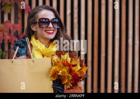 Bonjour novembre. heureuse femme moderne d'âge moyen en trench-coat brun avec sac à provisions et feuilles jaunes d'automne dans la ville. Banque D'Images