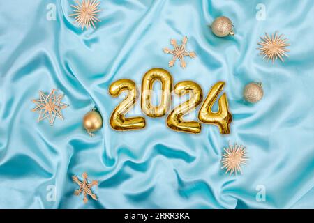 numéro 2024 écrit avec des ballons gonflables dorés à côté des décorations d'arbre de noël sur tissu satiné turquoise Banque D'Images