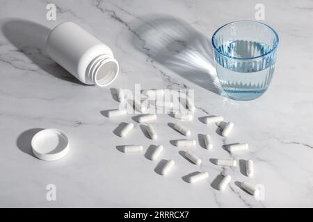 Pilules médicales blanches et verre d'eau sur un fond de table en marbre gris. Nature morte avec des ombres dures. Un tas de capsules éparpillées. Soins de santé conc Banque D'Images