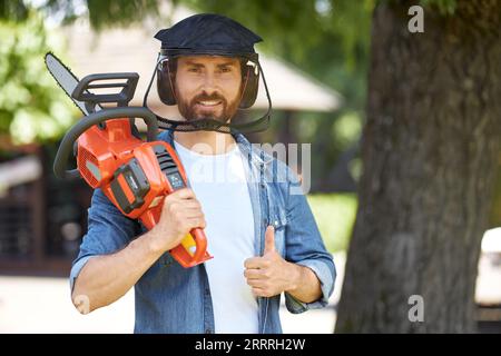 Travailleur barbu dans un écran de protection montrant le pouce vers le haut, tout en portant la tronçonneuse sans fil sur l'épaule à l'extérieur. Portrait de bricoleur souriant avec scie électrique moderne gestuant. Concept de jardinage. Banque D'Images