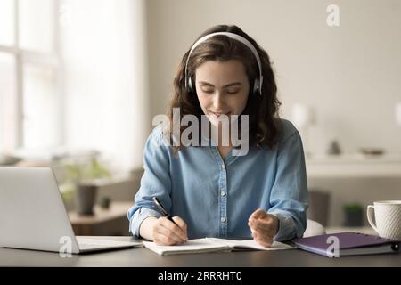 Fille étudiante en ligne positive et travailleuse dans des écouteurs écrivant des notes récapitulatives Banque D'Images