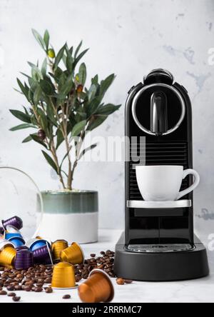 Tasse à café avec machine à expresso maison, plante verte et grains de café à arôme cru avec capsules dosettes dans le fond de cuisine clair. Banque D'Images