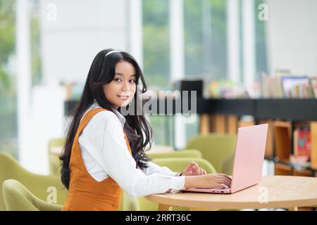 Belle adolescente asiatique avec ordinateur portable. Jeune femme avec les cheveux longs travaillant sur l'ordinateur dans le restaurant. Étudiant faisant ses devoirs. Banque D'Images